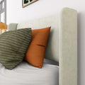Close-up of beige upholstered headboard and modern bedroom decor with green striped and rust-orange throw pillows on white bedding, bedside lamp and window