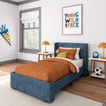 Modern kids bedroom decor with a blue upholstered twin bed, orange quilted bedding and pillows, white nightstands with orange lamps, framed "Young Wild and Free" wall art, pennant, plush lion and soccer ball