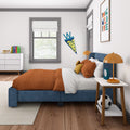 Modern kids bedroom interior with blue upholstered bed and orange bedding, stuffed lion, soccer ball, white dresser with books, orange bedside lamps, teal alarm clock, window with gray trim, colorful "Young Wild and Free" wall art and personalized "James" pennant