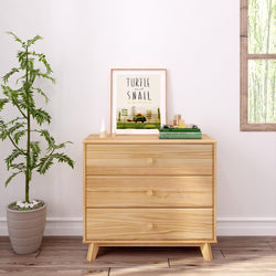 Wooden dresser with three drawers next to a potted plant in a bright room.