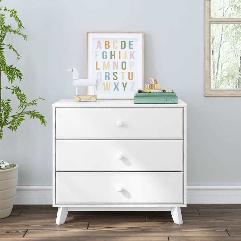 White three-drawer nursery dresser with pastel alphabet wall art, wooden pull toy horse, stacked children's books and alphabet blocks on top in a bright modern kids' room with potted plant and window