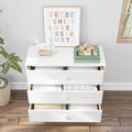 White nursery dresser with three open drawers filled with folded baby clothes, framed alphabet print, wooden pull toy and children's books on top, potted plant and hardwood floor