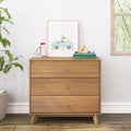 Mid-century modern three-drawer wooden dresser in a bright nursery, styled with a framed blue van print, toy sailboat, alphabet blocks, stacked books, potted plant and hardwood floor
