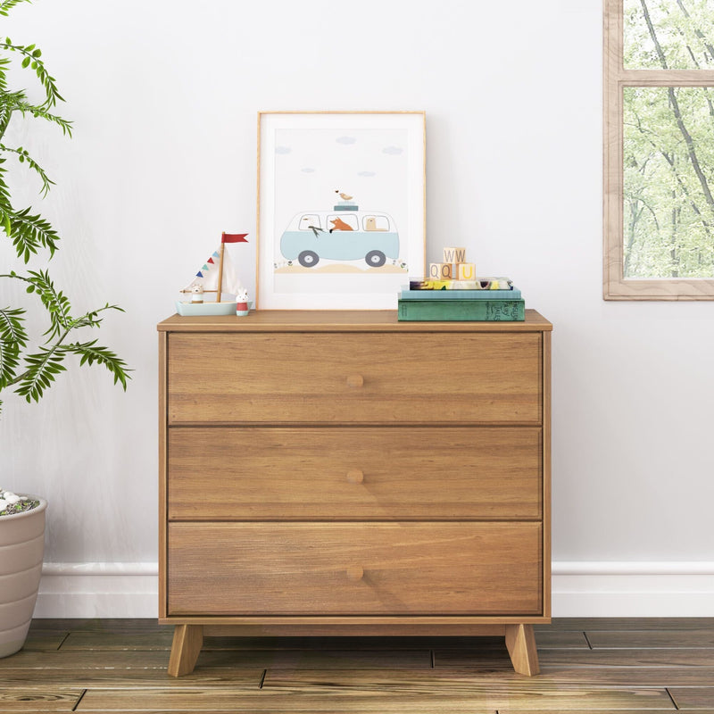 Mid-century modern three-drawer wooden dresser in a bright nursery, styled with a framed blue van print, toy sailboat, alphabet blocks, stacked books, potted plant and hardwood floor