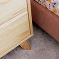 Close-up of light pine wooden nightstand leg on beige carpet beside a pink upholstered bed frame with floral bedding