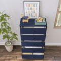 Navy blue five-drawer nursery dresser with open drawers showing folded baby clothes, wooden toy horse, framed alphabet print, children's books and potted plant on hardwood floor