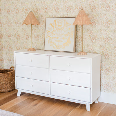 White six-drawer dresser styled with framed gold butterfly art, wicker table lamps and floral wallpaper for nursery or boho kids' room decor
