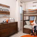 Young boy in modern kids bedroom with dark wooden bunk bed, checkered bedding, wooden dresser with soccer and basketball, orange accents and "dream" wall art