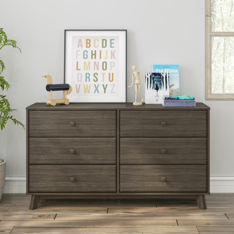 Modern wooden dresser with six drawers, decorated with colorful framed alphabet art, books, wooden duck toy, and mannequin on top, in bright room with window and plant.