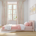 Pastel pink toddler bed with smiley sun pillow and patterned blanket, pink-and-cream striped curtains framing a wooden window in a bright nursery
