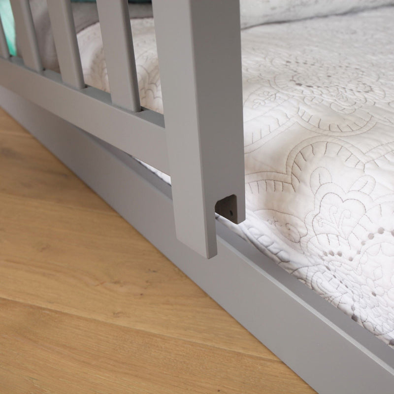 Gray crib rail and white quilted bedding on wooden floor.