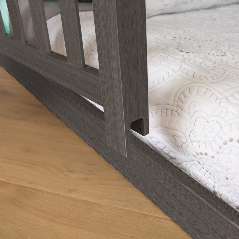 Wooden crib detail with white quilted bedding on a light wood floor.
