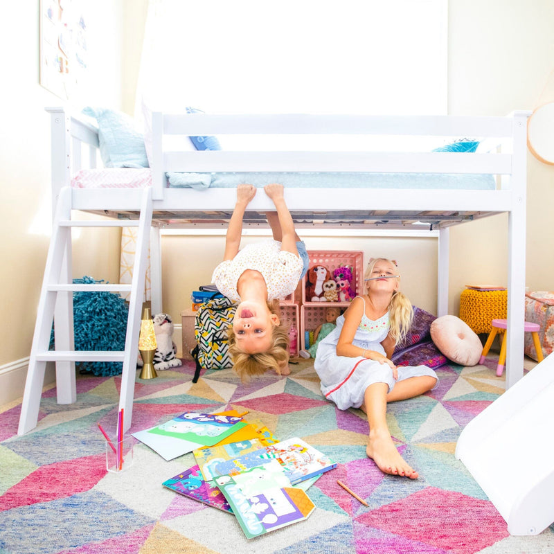 Two young girls playing under a white loft bed in a colorful kids' bedroom with children's books and toys on a geometric rug