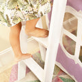 Bare feet stepping on a white wooden ladder beside a tropical floral dress and pastel purple fabric — nursery decor detail for kids' room, white ladder and cozy play area styling