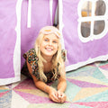 Smiling blonde girl in floral dress peeking out of a purple indoor play tent with a sleep mask on her forehead while lying on a colorful geometric rug