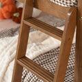 Close-up of a natural wood bunk bed ladder with grooved steps beside cozy cream blanket, gingham plaid bedding, and autumn pumpkin decor