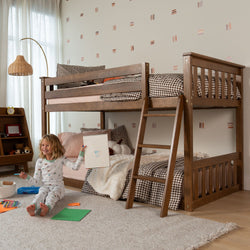 Wooden twin-over-twin bunk bed in cozy children's bedroom with ladder, gingham bedding, plush rug, bedside shelf and child drawing on floor with art supplies