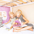 Two young girls reading a colorful children's book together in a cozy under-bed fort reading nook beneath a bunk bed — playful bedtime storytime for kids