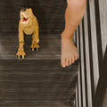 Toy T-Rex dinosaur on dark wooden stairs next to a bare foot, close-up indoor play scene