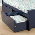 Navy blue wooden daybed with open under-bed storage drawer revealing teal and gray patterned pillow and navy blanket on light hardwood floor
