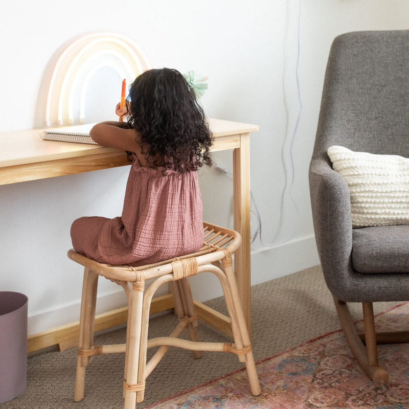 Young girl drawing at a light wood desk while seated on a rattan stool in a cozy nursery with a rainbow lamp, gray rocking chair and textured rug