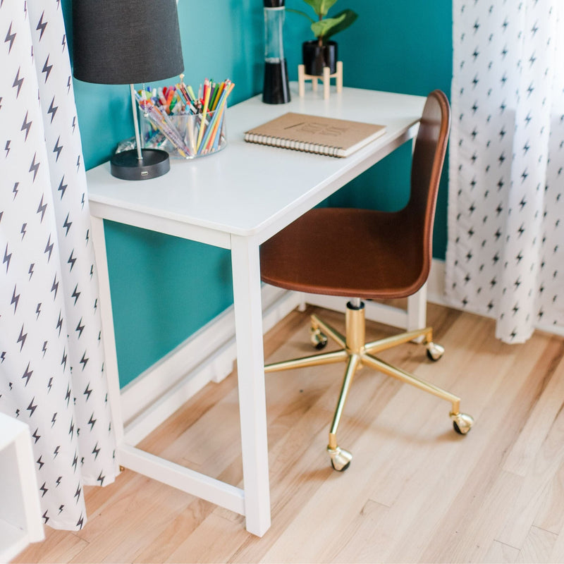 Modern white desk home office setup with brown swivel chair (gold base), black lamp, notebook and colored pencils against teal accent wall