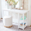 White vanity table with scalloped mirror, striped round ottoman, pastel floral wallpaper, fresh flower vases and children's books in a bright kids' room