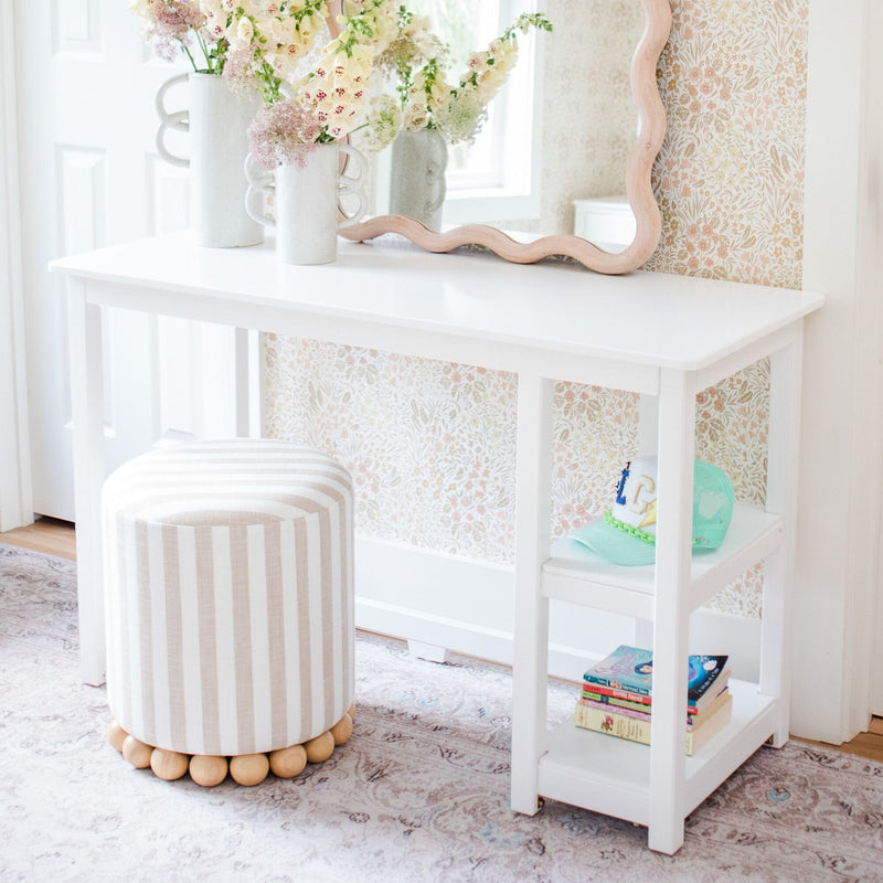 White vanity table with scalloped mirror, striped round ottoman, pastel floral wallpaper, fresh flower vases and children's books in a bright kids' room