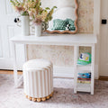 White vanity desk with scalloped mirror and striped round ottoman against floral wallpaper, styled with vases, books and a green cap — kids bedroom decor