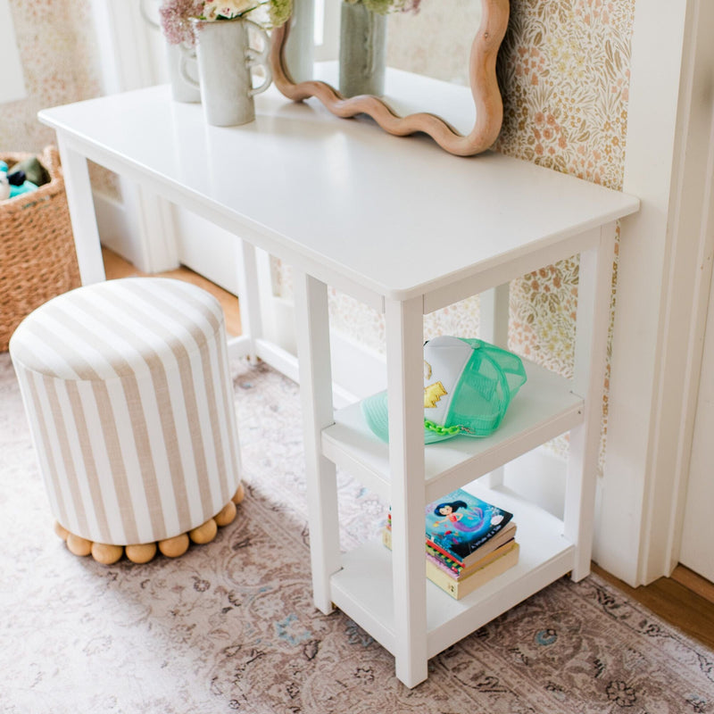 White children's vanity desk with scalloped mirror, striped round ottoman, mint green trucker hat and stacked children's books on lower shelves styled on a floral nursery rug