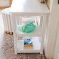 White bedside table with a mint green mesh trucker hat on the middle shelf and stacked children's books on the lower shelf beside a striped ottoman and patterned rug, cozy nursery decor