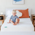 Toddler mattress: smiling toddler sitting on a white breathable memory foam mattress in a bright modern nursery with a cloud-shaped pillow and terracotta cushion