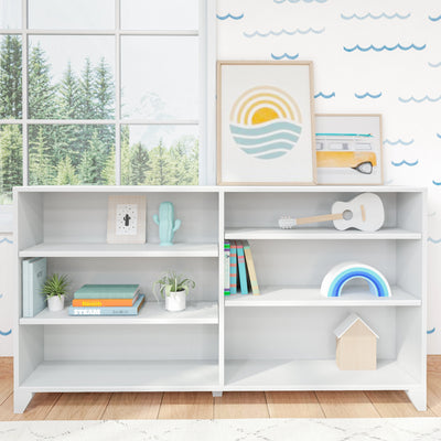 Bright modern nursery shelf with framed sun-and-waves print, yellow toy van, white ukulele, pastel rainbow toy, turquoise cactus figurine, children's books and potted plants on white shelving by a large window overlooking evergreen trees, minimalist kids room decor.