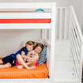 Two smiling children cuddling on the lower bunk of a modern white bunk bed with orange quilt, patterned pillows and built-in stairs in a bright kids' bedroom