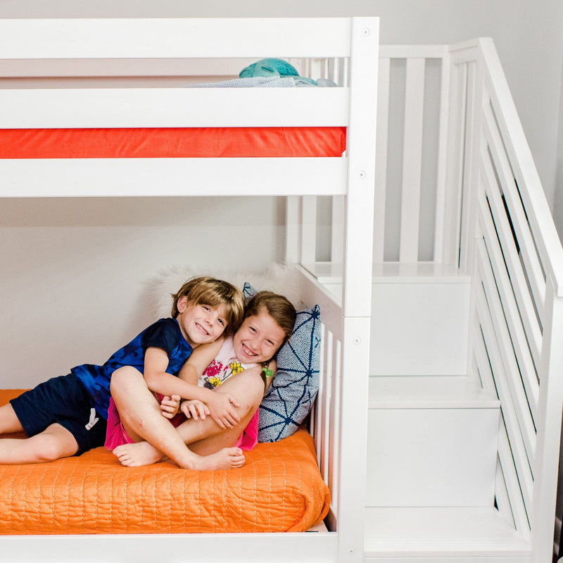 Two smiling children cuddling on the lower bunk of a modern white bunk bed with orange quilt, patterned pillows and built-in stairs in a bright kids' bedroom