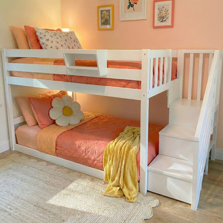 White wooden bunk bed with staircase, coral-pink bedding, daisy accent pillow and yellow throw in a pastel pink kids' bedroom with floral wall art — space-saving girls' bunk bed and kids room decor