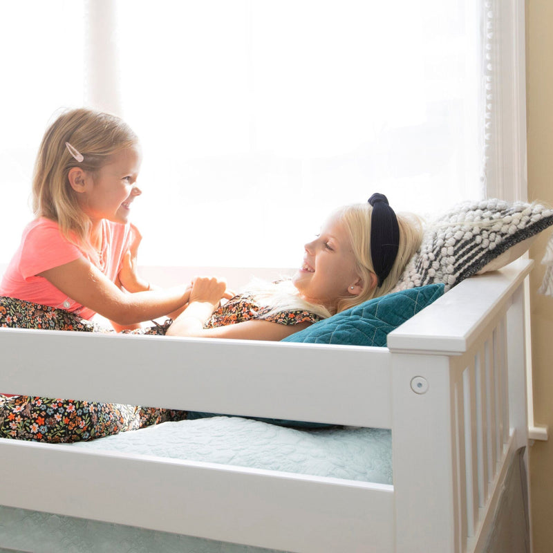 Two young girls smiling and playing on a cozy white bed near a sunlit window.