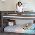 Smiling child standing on a gray wooden kids bunk bed in a bright children's bedroom with pastel patterned bedding, playful pillows and farmhouse-style wall art