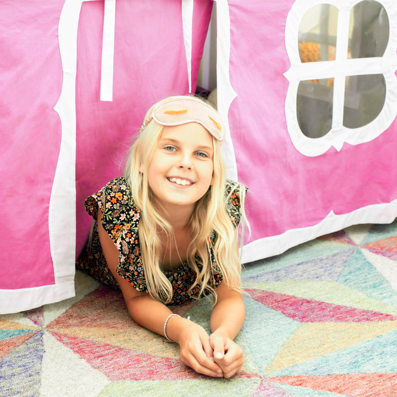 Young girl with a sleep mask smiling in a decorated play tent on a colorful rug.