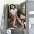 Smiling child sitting on wooden bunk bed stairs in a cozy neutral kids bedroom with a stuffed rabbit toy and leather backpack