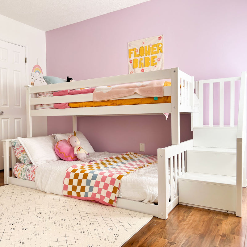 White wooden bunk bed with staircase in pink children's bedroom, colorful checkered blanket, unicorn plush toy and "Flower Babe" wall art
