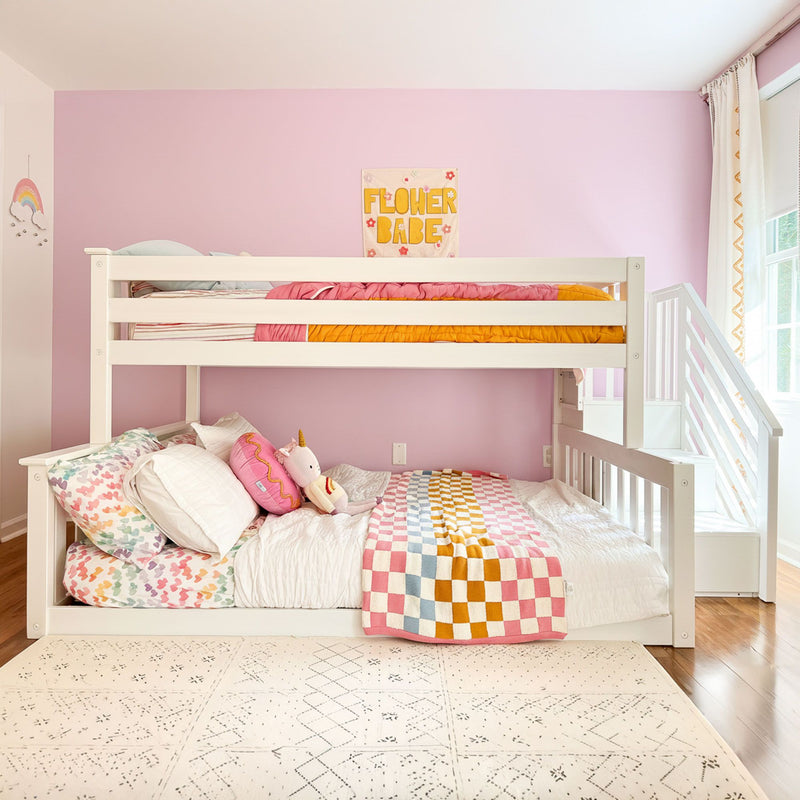 White wooden bunk bed with stairs in pink girls' bedroom, colorful checkered quilt, heart-print pillows, unicorn plush and "Flower Babe" wall art