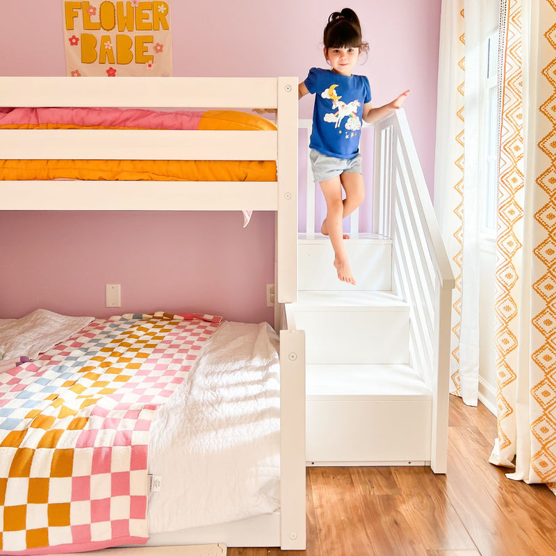 White bunk bed with stairs in pink kids' bedroom, colorful checkered quilt, hardwood floor and young girl stepping down the staircase