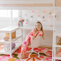 Smiling girl in pink tie-dye pajamas seated at a white desk chair in a bright girls' bedroom study nook with fairy lights, floral rug, and loft bed