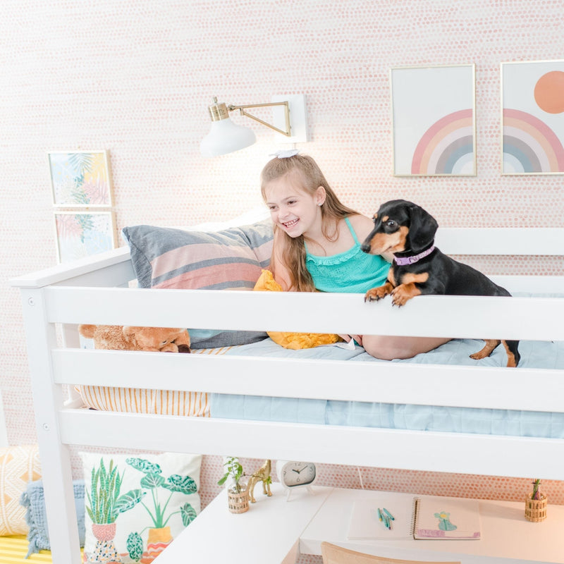 Smiling young girl on a white loft bed with a black-and-tan dachshund in a pastel kids' bedroom featuring rainbow wall art, patterned pillows, and cozy decor.