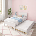 White wooden single bed with pull-out trundle in pastel girls' bedroom featuring pink scallop wallpaper, rainbow and sun wall art, blue throw and bedside lamp