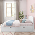 Sunlit pastel pink kids' bedroom with white storage twin bed, blue throw blanket, pastel pillows, geometric bedside lamp, potted plant and rainbow wall art