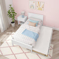 Modern kids' bedroom with white pull-out trundle bed, pink scallop wallpaper, pastel bedding, white nightstand, potted plant and rainbow sun wall art