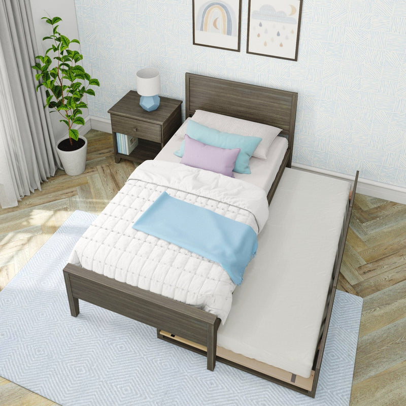 Gray wooden twin bed with pull-out trundle, white quilt, blue throw and pastel pillows, nightstand lamp and potted plant