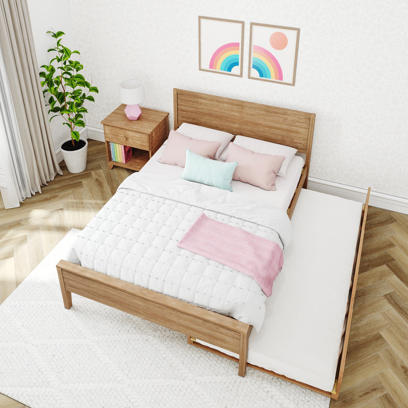 Cozy pastel bedroom with wooden bed and pull-out trundle, white quilt and pink throw, pastel pillows, wooden nightstand with lamp and books, potted plant and rainbow wall art
